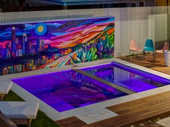 Nighttime backyard plunge pool glowing purple with LED lights, modern wooden deck and pastel chairs beside a large colorful urban skyline and desert-flora mural on a corrugated fence.