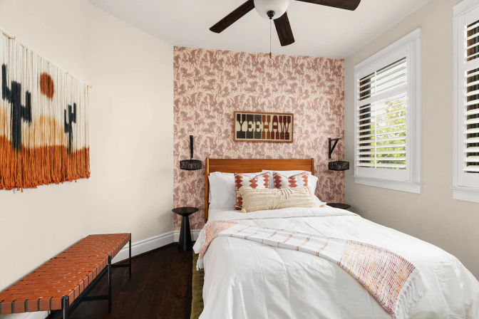 Sunlit boho bedroom with wooden bed and white bedding against a blush floral accent wall, patterned pillows, woven wall hanging, leather bench, dark hardwood floor and plantation shutters.