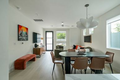 Bright modern open-plan living and dining room with a black oval table and six brown chairs, sculptural white pendant light, cozy sofa with colorful pillows, wall-mounted TV, wood sideboard, red velvet bench, light wood floors and large windows letting in natural light.