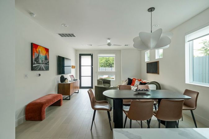 Bright modern open-plan living and dining room with a black oval table and six brown chairs, sculptural white pendant light, cozy sofa with colorful pillows, wall-mounted TV, wood sideboard, red velvet bench, light wood floors and large windows letting in natural light.