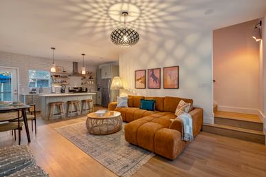 Cozy modern open-concept living room and kitchen with warm wood floors, a tufted caramel sectional, woven round coffee table, island with three bar stools, pendant lights casting decorative shadows, neutral cabinetry and framed artwork.
