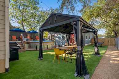 Inviting suburban backyard with black gazebo covering a wooden dining table and mixed chairs on artificial turf, raised above-ground pool with blue umbrellas and orange lounge chairs, string lights and fenced yard.