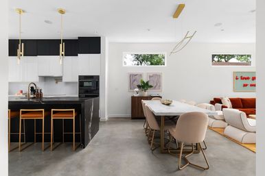 Sunlit modern open-concept kitchen, dining and living room with black marble island, gold-accent bar stools and pendants, marble dining table with blush chairs, concrete floors and rust sofa.