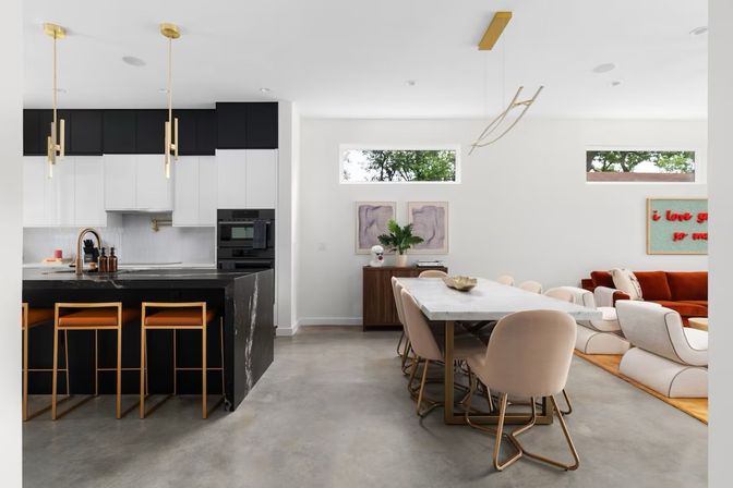 Sunlit modern open-concept kitchen, dining and living room with black marble island, gold-accent bar stools and pendants, marble dining table with blush chairs, concrete floors and rust sofa.
