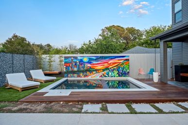 Modern backyard plunge pool with wooden deck and reflective water, two white lounge chairs on artificial turf, vibrant sunset city skyline mural on corrugated fence, colorful patio chairs and greenery under a clear blue sky.