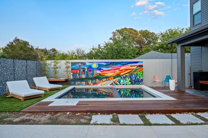 Modern backyard plunge pool with wooden deck and reflective water, two white lounge chairs on artificial turf, vibrant sunset city skyline mural on corrugated fence, colorful patio chairs and greenery under a clear blue sky.