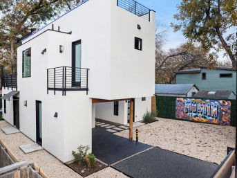 Modern white two-story home with rooftop deck and balcony, gravel yard and driveway, colorful “Welcome to Austin, Texas” mural on a neighboring wall and leafy trees