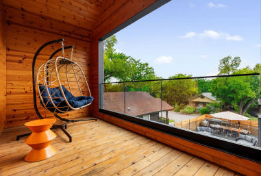 Cozy wooden balcony with rattan hanging chair and navy cushions, bright orange side table, glass railing and a tree-lined neighborhood view with backyard patios.
