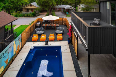 Modern Austin backyard with a rectangular blue pool and floating lounger, string lights overhead, orange beanbag loungers and gray cushioned seats on a dark wood deck, dining table under an umbrella, a muraled wooden privacy fence, and a black second-story deck.