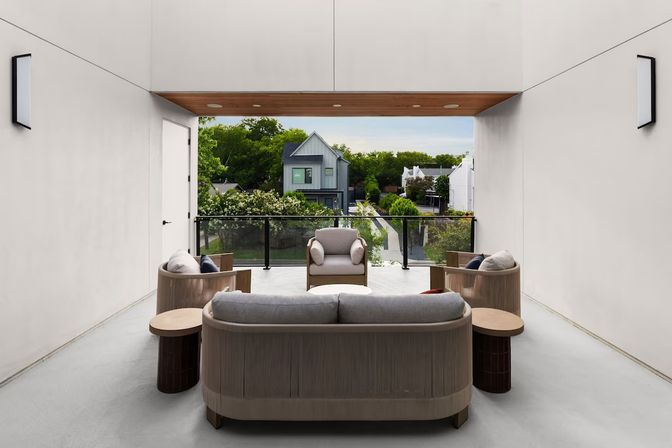 Modern covered balcony lounge with curved neutral sofa and armchairs around small tables, wood-paneled ceiling and glass railing overlooking a tree-lined suburban street and contemporary houses.