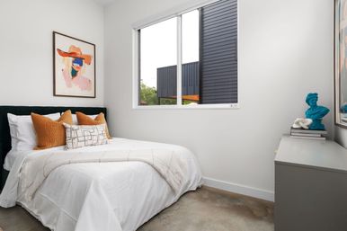 Cozy modern minimalist bedroom interior with white bedding, rust-orange accent pillows, abstract portrait print above the bed, concrete floor, large window with daylight, and a gray dresser topped with a blue bust sculpture.