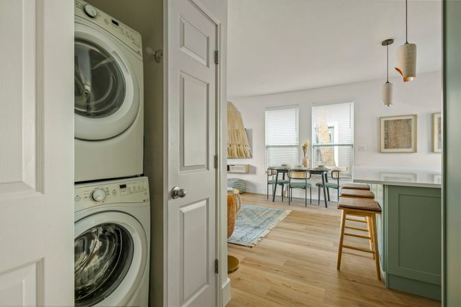 Peek into a bright modern apartment: stacked washer/dryer in a closet opening to an open-plan living area with a mint-green breakfast bar, wooden stools, dining table by sunlit windows and light hardwood floors.