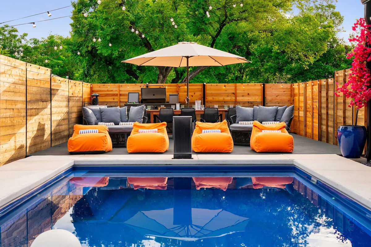 Backyard swimming pool with blue water reflecting a large patio umbrella; wooden deck with bright orange bean‑bag loungers, gray cushioned sofas, dining table and grill, string lights and lush green trees