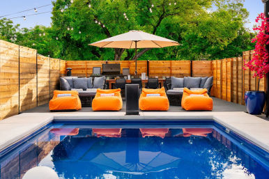 Backyard swimming pool with blue water reflecting a large patio umbrella; wooden deck with bright orange bean‑bag loungers, gray cushioned sofas, dining table and grill, string lights and lush green trees