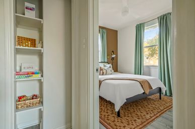Sunny guest bedroom with white bedding and brown throw on a simple bed, mint-green curtains, woven area rug, bedside reading lamp, and built-in shelving stocked with classic board games like Monopoly, Connect Four and Uno.