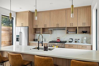 Bright modern kitchen with light wood cabinets and marble-look quartz surfaces, large white island with undermount sink and black spring-coil faucet, stainless refrigerator and range with red kettle, three tan leather bar stools and brass pendant lights.