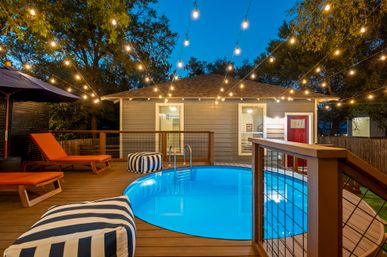 Cozy evening backyard pool deck with a glowing round above-ground pool on a wooden deck, striped poufs, orange lounge chairs, overhead string lights and a small gray cottage with a red door.
