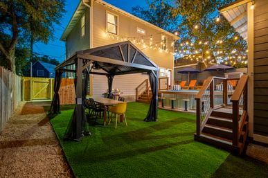 Evening residential backyard patio with a covered gazebo and dining table on artificial turf, warm string lights overhead, raised deck with above-ground pool and orange lounge chairs under umbrellas.