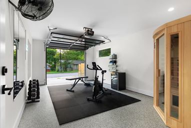 Bright garage home gym with exercise bike, adjustable bench, dumbbell rack on black rubber mats, wooden sauna, mini fridge and open roll-up door showing suburban street and trees.