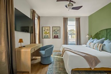 Sunlit modern bedroom with green velvet arch headboard, king bed dressed in blue pillows and striped bolster, wooden desk with small lamp, blue swivel chair, wall-mounted TV, two whimsical framed prints, beige curtains and ceiling fan.