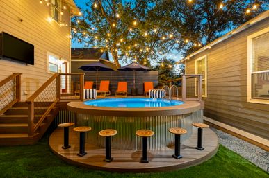 Cozy backyard patio at dusk featuring a round above‑ground pool with wooden deck stools, orange lounge chairs and navy umbrellas, warm string lights strung through trees and an inviting outdoor seating area.
