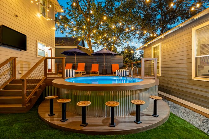 Cozy backyard patio at dusk featuring a round above‑ground pool with wooden deck stools, orange lounge chairs and navy umbrellas, warm string lights strung through trees and an inviting outdoor seating area.