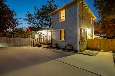 Cozy twilight view of a two-story gray house with warm-lit windows, a small porch with steps, planters and a wooden fence beside a wide concrete driveway