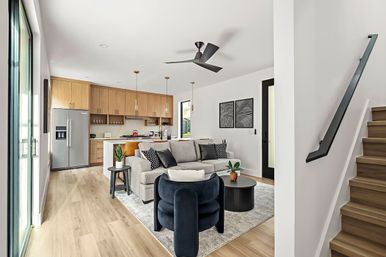 Sunlit modern open-plan living room and kitchen with light wood floors, gray sofa, navy accent chair, round coffee table, island with pendant lights, stainless fridge, sliding glass doors and wooden staircase.