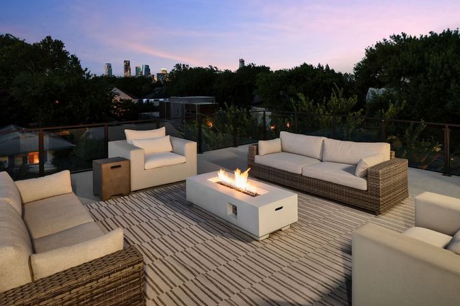 Cozy rooftop patio at dusk with modern wicker sofas and cushioned armchairs arranged around a rectangular fire pit, striped outdoor rug and glass railing with city skyline beyond.