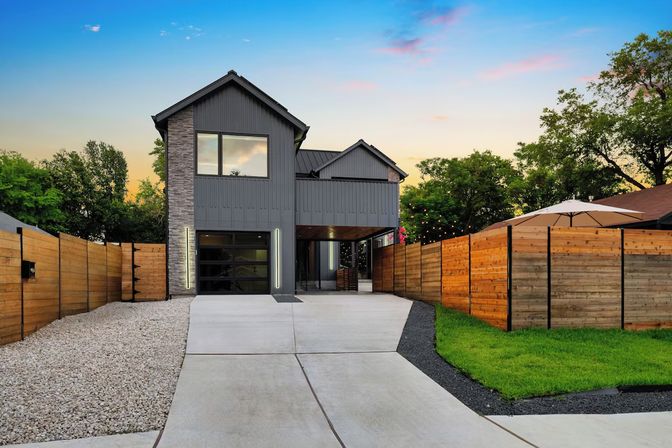 Sleek two-story modern home with gray siding and stone accent, glass garage door and wide concrete driveway flanked by wood privacy fences, gravel bed and green lawn, patio umbrella and string lights under a colorful sunset sky.