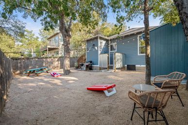 Sunny suburban backyard with blue-gray house, string lights hanging between mature trees, gravel ground with picnic table, cornhole boards, wicker bistro seating and a barbecue for casual outdoor entertaining.