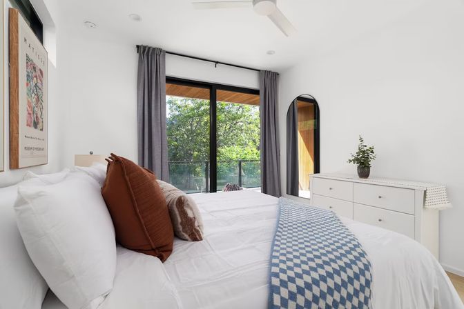 Bright modern minimalist bedroom with white bedding, rust and patterned throw pillows, blue check blanket, sliding glass doors to a balcony with leafy treetop view, gray curtains, white dresser with potted plant and arched mirror, and ceiling fan.
