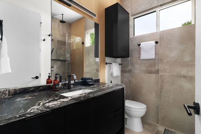 Sleek modern bathroom with black vanity and marbled countertop, wall mirror, glass walk-in shower, beige large-format tile walls, white toilet, towel racks and a narrow window providing natural light.