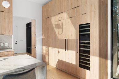 Sunlit modern kitchen with floor-to-ceiling light wood cabinets, integrated wine fridge, marble island, and large window.