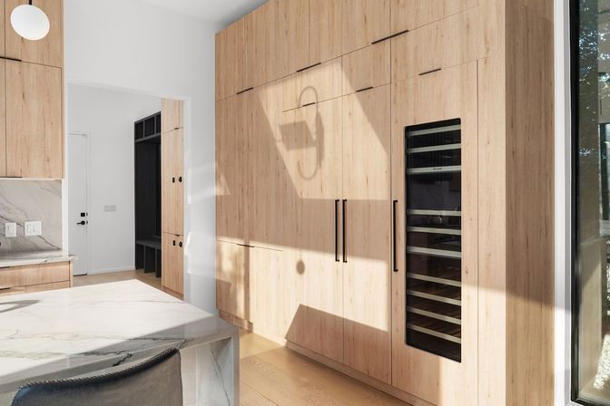 Sunlit modern kitchen with floor-to-ceiling light wood cabinets, integrated wine fridge, marble island, and large window.