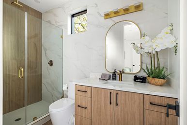 Bright contemporary residential bathroom with glass shower and brass hardware, wood-tone vanity with white marble-look countertop, gold arched mirror, white orchids and a gold faucet.
