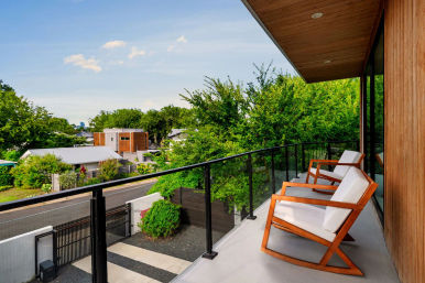 Inviting modern wood-clad balcony with glass railing and two cushioned wooden lounge chairs overlooking a leafy suburban street and contemporary homes under a bright blue sky.