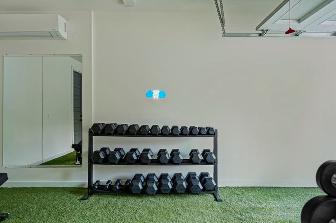 Bright garage-style home gym with a three-tier rack of black hex dumbbells on green turf, mirrored wall, wall-mounted AC, and open garage door track — ready for a workout.
