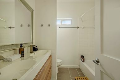 Bright modern minimalist bathroom with double-sink vanity on a wood cabinet, large framed mirror, folded towel and soap dispenser on the counter, white tiled bathtub-shower with toiletries on the tub edge, small high window and gray ceramic floor tiles.