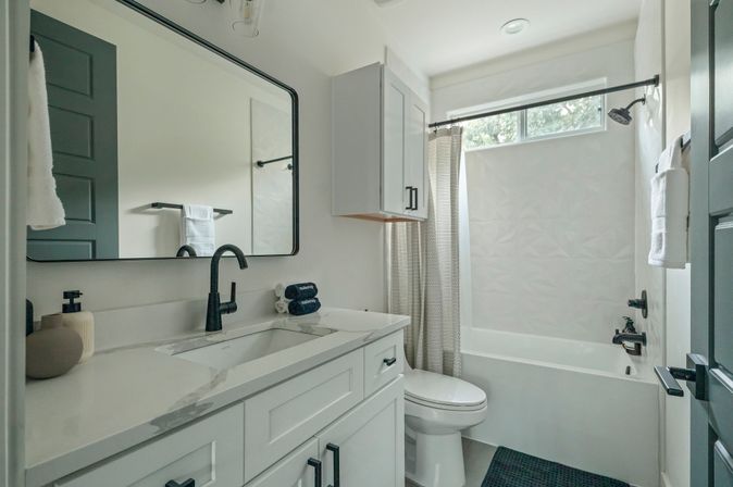 Bright modern residential bathroom with white vanity and marble-look countertop, black matte faucet and hardware, large framed mirror, shower-tub with high window and patterned curtain, wall cabinet and neatly folded towels.