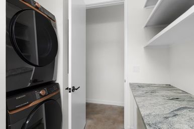Compact modern laundry room with stacked front-load washer and dryer, white open shelving, stone-look countertop, concrete floor and open door.