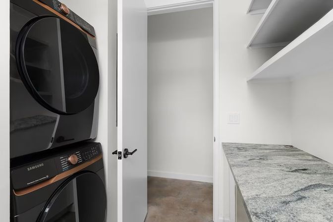 Compact modern laundry room with stacked front-load washer and dryer, white open shelving, stone-look countertop, concrete floor and open door.