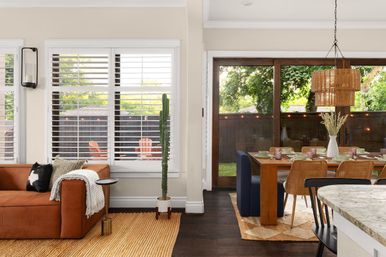 Sunlit open-plan living room and dining area with caramel leather sofa, tall cactus, wooden dining table, rattan pendant light, and sliding glass doors to a backyard patio with orange Adirondack chairs.
