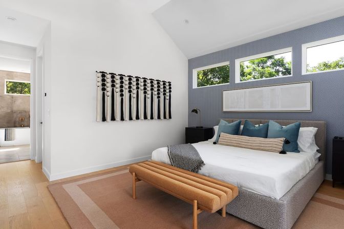 Bright modern master bedroom with blue chevron accent wall, low upholstered bed with teal pillows, tan bench, black-and-white tassel wall hanging and three narrow windows overlooking leafy trees