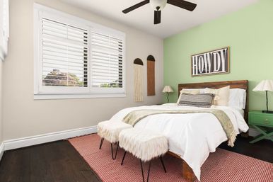 Cozy modern bedroom interior with mint green accent wall, wooden bed with white linens and striped pillow, 'HOWDY' wall art, two fluffy ottomans, plantation shutters, dark hardwood floors and a green nightstand.