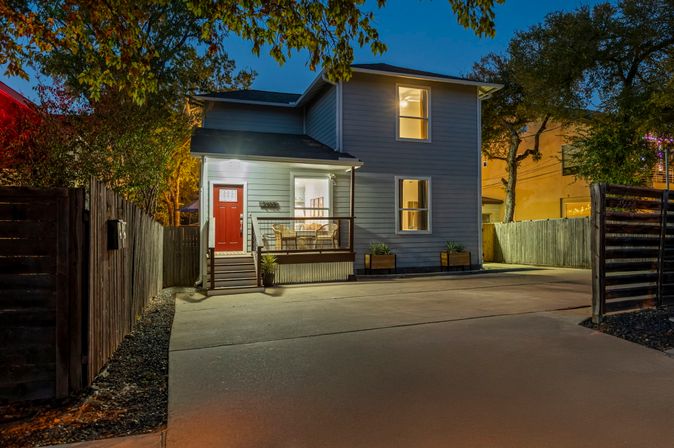 Cozy two-story home exterior at dusk — red front door, lit porch with wicker chairs, concrete driveway, planters and wooden privacy fence in a residential neighborhood.