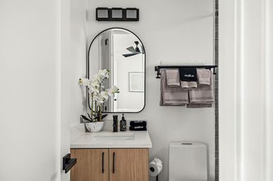 Crisp modern bathroom with oak vanity and white countertop, black-framed arched mirror, potted white orchids, matte black faucet and soap, gray folded towels on a wall rack, and a compact white toilet