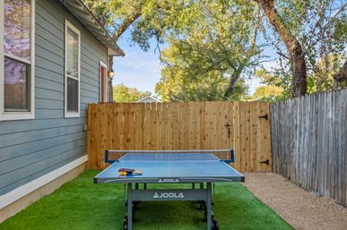 Sunny fenced backyard with an outdoor table tennis (ping pong) table, paddles and balls on a blue JOOLA table set on green artificial turf beside a blue-sided house and wooden fence.