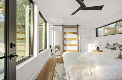 Bright modern bedroom with large windows overlooking trees, white walls, hardwood floors, king bed with textured white bedding and throw, wooden sliding barn door, black ceiling fan and bedside lamp — sunlit minimalist design.