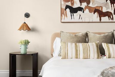 Cozy modern bedroom interior with beige upholstered headboard, white bedding, striped lumbar pillows and velvet cushions, framed horse artwork above the bed, rattan wall sconce, and a round black bedside table holding a mint vase with flowers.